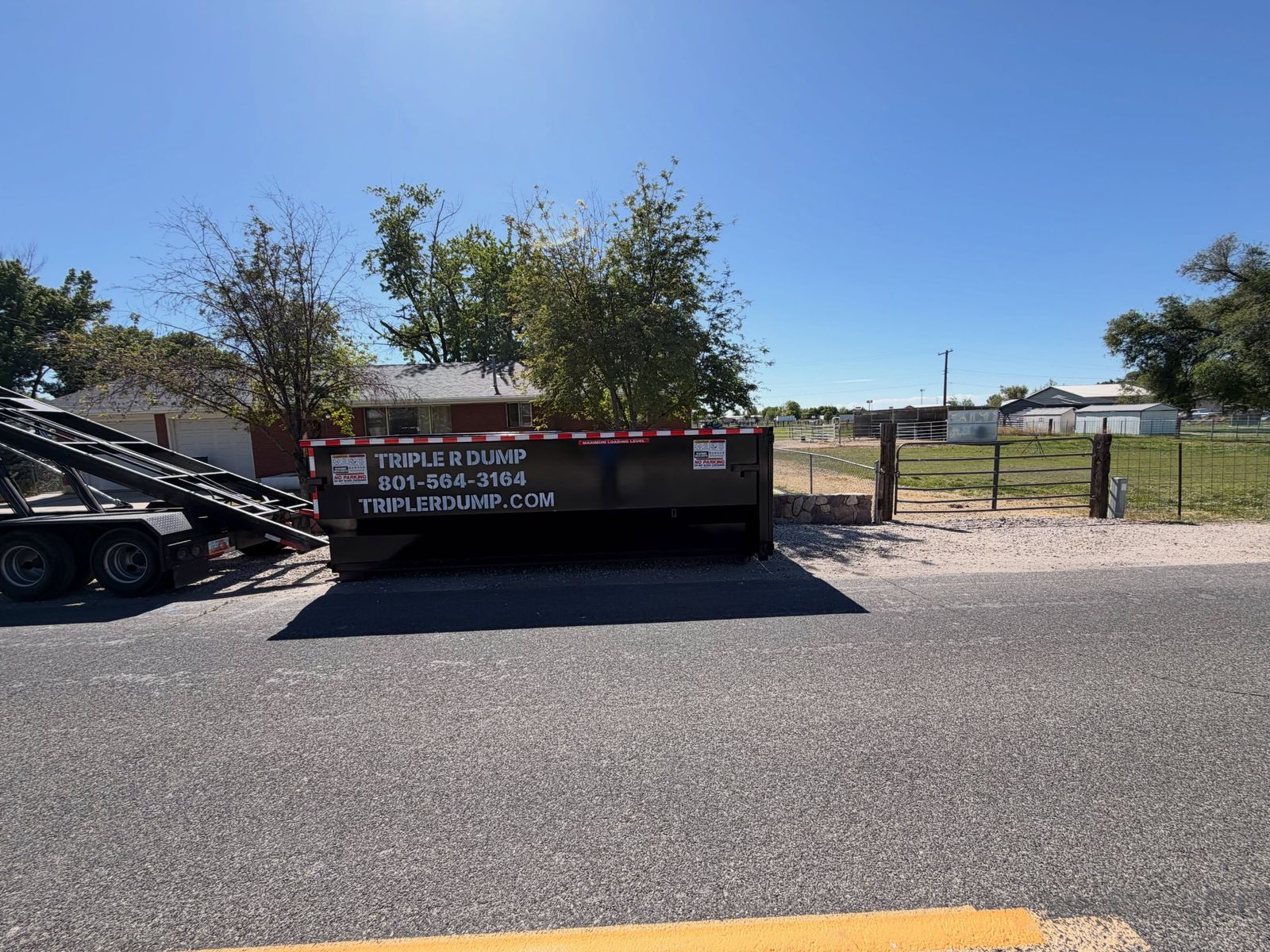 Triple R Dump roll-off dumpster dropped at a curb beside a fenced yard, roll-off trailer behind it